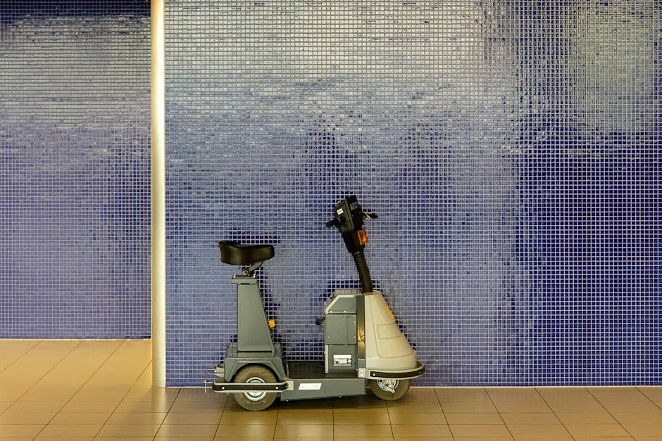 A small electric mobility scooter with a black cushioned seat and a grey body is situated on a light brown tiled floor, positioned against a large wall covered in small, square, blue mosaic tiles. The scooter has a black handlebar with controls and a small orange reflector beneath the handlebars. The environment appears to be an indoor space, possibly within a commercial or public building, with a vertical white pole visible on the left side of the image. The blue mosaic-tiled wall provides a textured backdrop, and there are no other objects or furnishings visible in the scene. The setting suggests a location where accessible transportation or mobility aids are stored or used, subtly reflecting the context of managing access and mobility within a waste removal or facility area, as associated with private waste handling services like Waste Removal Holland Park.