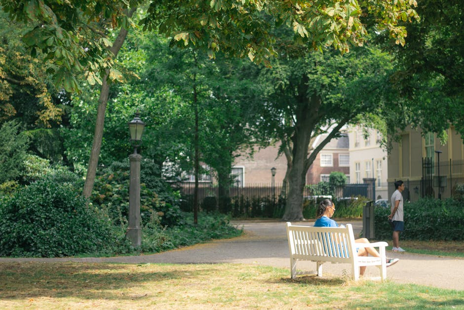 A peaceful urban park scene featuring a white wooden bench placed on a paved pathway, with a young woman seated on it wearing a blue top and light-colored shorts, appearing to be engaged in her smartphone. Nearby, a young boy wearing a white t-shirt and dark shorts stands on the pathway, looking towards the distance. The park is lush with greenery, including large trees with broad, green leaves and dense shrubbery on the left side, providing shade and a natural environment. A traditional-style lamppost is situated behind the bushes to the left of the bench, contributing to the park’s classic ambiance. In the background, there are residential buildings with light-colored facades and multiple windows, partially obscured by the trees and a black wrought-iron fence. The scene is illuminated by bright, natural daylight, creating a calm and inviting atmosphere, typical of a space where residents might engage in walking, relaxing, or informal outdoor activities, reflecting the context of private or alternative waste disposal and the natural setting around local rubbish removal services in an urban environment.