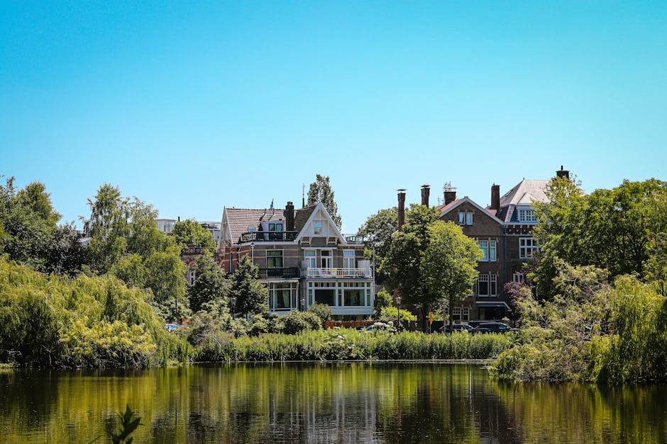 A row of residential buildings situated behind a lush, green park landscape with trees and bushes along the water's edge. The buildings are multi-storey, featuring a mix of brick and painted facades, with large windows, balconies, and pitched roofs. The scene is illuminated by natural daylight under a clear, bright blue sky. The foreground shows a calm body of water reflecting the greenery and architecture, with a narrow strip of grassy banks and overhanging branches. This setting illustrates the environment where private waste collection or rubbish removal services might take place, highlighting the proximity of residential properties to natural surroundings, useful for context in an article about alternative waste handling options provided by Waste Removal Holland Park.