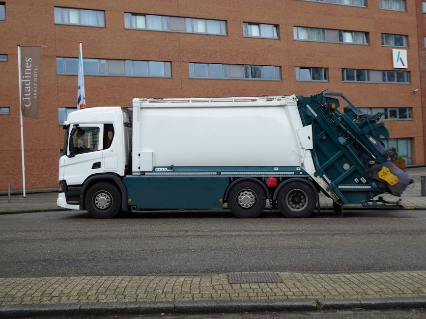 A large white and dark blue rubbish collection truck is parked on a city street in front of a modern red-brick building with multiple rectangular windows. The vehicle features a compact cab with a side mirror and a front windshield, and extends to a sizeable rear loading compartment. The rear section is equipped with mechanical arms and a hydraulic lifting mechanism, primarily painted dark blue with white accents, designed for waste collection and disposal. The truck is positioned adjacent to a paved sidewalk with patterned brickwork, and the background includes a flagpole with a flag and a vertical sign indicating nearby accommodation. The environment appears to be during daylight, with soft natural light reflecting off the vehicle’s surfaces, typically used for private waste handling and alternative rubbish collection services in urban areas like Holland Park.