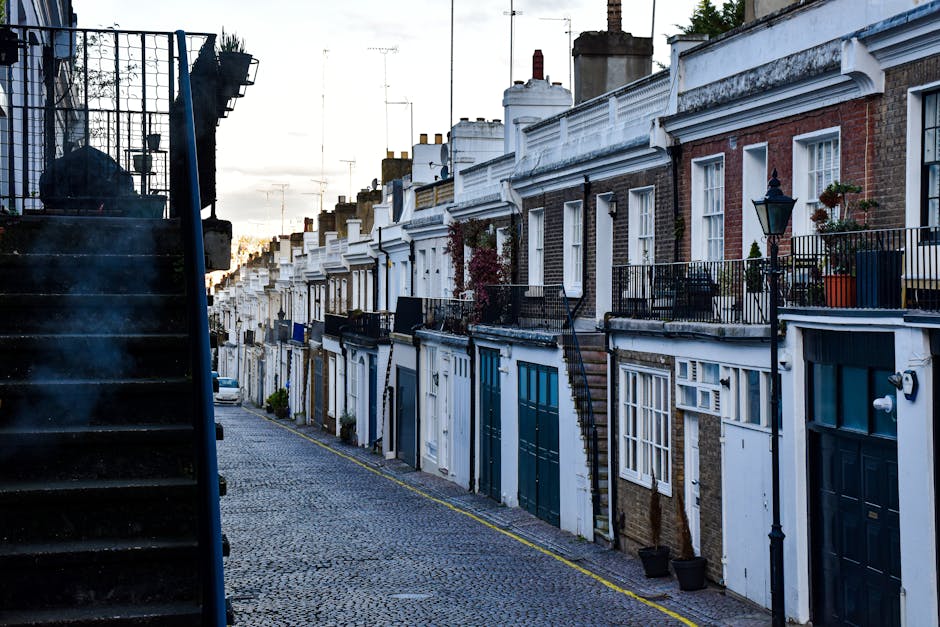 The image depicts a narrow residential cobblestone street lined with rows of terraced brick houses featuring white and dark blue doors and window frames. Each property has a small front terrace or balcony, some decorated with potted plants and outdoor furniture, with black wrought iron railings and street lamps mounted on the facades. On the left side of the image, a set of black exterior stairs with a metal railing ascends from a lower level, partially obscuring some of the view. The sky in the background suggests an early evening or late afternoon setting, with soft natural light illuminating the scene and casting gentle shadows. The environment appears quiet and well-maintained, characteristic of an urban area where private waste handling or independent rubbish collection services, such as those offered by Waste Removal Holland Park, might be utilized for on-site rubbish clearance or disposal outside of municipal collection. The overall atmosphere is tidy, with a focus on the architectural details of the row of terraced houses and the street’s narrow, pedestrian-friendly layout.