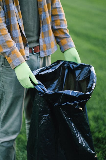 A person wearing a plaid shirt with yellow, gray, and blue checkered patterns, along with green protective gloves and gray trousers, is holding open a large black rubbish bag made of thick, shiny plastic material. The bag is partially filled and is positioned outdoors on a grassy area, with the surrounding environment featuring lush green grass and blurred background foliage. The individual appears to be preparing the rubbish bag for waste collection, which could relate to private waste handling or on-site clearance services offered by Waste Removal Holland Park, emphasizing the importance of proper waste disposal and rubbish removal practices for maintaining clean outdoor spaces.