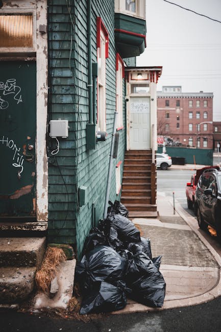 A row of black garbage bags filled with waste are piled against the side of a building with green, weathered wooden siding and red window frames, situated on a sidewalk next to a set of wooden stairs leading up to a white door. The bags are placed on a concrete surface adjacent to a paved driveway, where a black vehicle is parked. In the background, there are other buildings and structures, indicating an urban environment. The scene appears to have natural daylight, with an overcast sky casting soft, diffuse light over the area. The image captures a typical example of private rubbish accumulation outside a residential or commercial property, aligning with waste management service needs for skip or rubbish removal providers like Waste Removal Holland Park, which may offer alternative waste handling options for such collection.