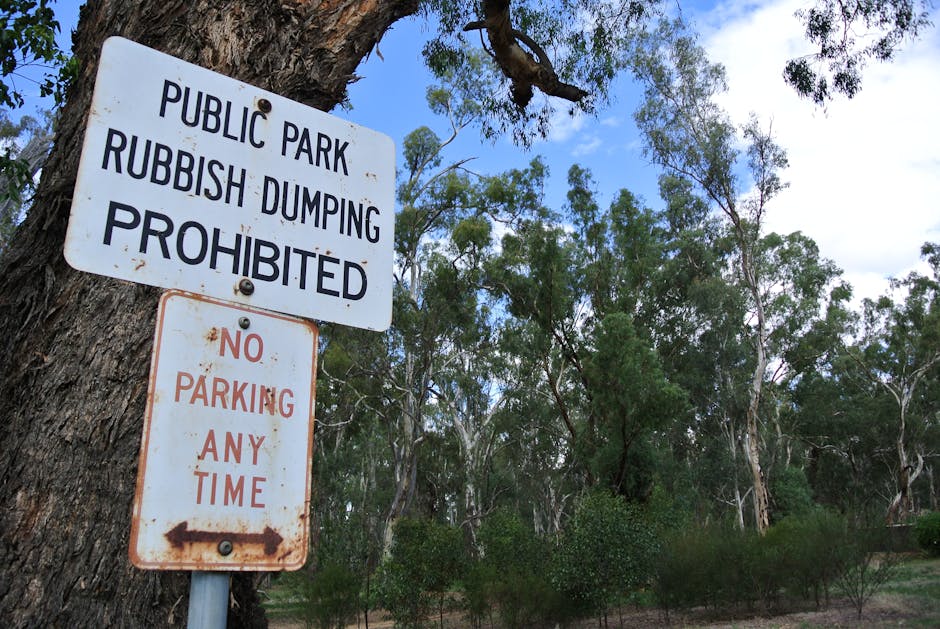 A pair of rectangular metal signs attached to a thick, textured tree trunk in a public park setting. The top sign features bold black text on a white background, stating 'PUBLIC PARK RUBBISH DUMPING PROHIBITED,' with visible screws securing it in place. Below, a smaller sign with a rusted appearance displays 'NO PARKING ANY TIME' in red text, with an arrow pointing to the left. The background includes numerous tall eucalyptus trees with slender, twisting trunks and dense green foliage, set against a partly cloudy sky with patches of blue. The scene suggests a natural environment where waste disposal is restricted, aligning with regulations similar to those outlined by local councils for private waste handling or on-site clearance, as managed by waste removal services such as Waste Removal Holland Park. The natural lighting highlights the textures of the bark and leaves, emphasizing the outdoor setting consistent with rubbish management and environmental protection practices.
