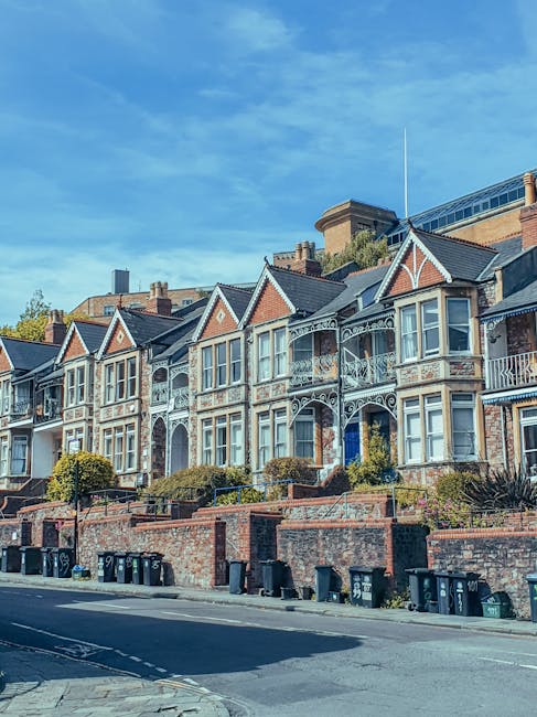 A row of Victorian-style terraced houses situated on an elevated street with a brick retaining wall and steps leading up to each property. The houses feature ornate white wrought-iron balconies and decorative trim on gabled roofs covered with dark slate tiles. Large bay windows with white frames dominate the front façades, and small front gardens with shrubs and plants are visible behind the railings. In the foreground, there are several black wheelie bins lined up along the sidewalk, suggesting waste collection or clearance activities. The background includes a modern multi-storey building with glass and brick elements under a clear blue sky with some wispy clouds, creating a contrast between historic and contemporary architectural styles. The scene appears to be during daylight hours with natural sunlight illuminating the area, providing a neutral and professional view of the residential street potentially linked to private waste management services such as those offered by Waste Removal Holland Park.